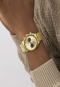 Gold-Chronograph-Uhr mit einem Metallarmband, goldenem Zifferblatt, schwarzen Nebenziffern und strukturierten Details, getragen an einem Handgelenk mit einem beigen Strickpullover.