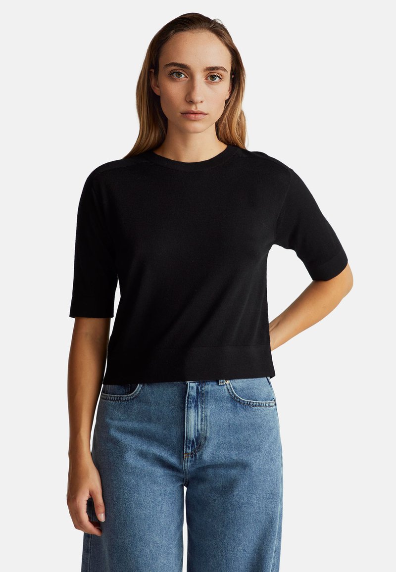 Młoda kobieta z długimi blond włosami, ubrana w czarny sweter z krótkim rękawem i niebieskie dżinsy z wysokim stanem, na jednolitym tle.