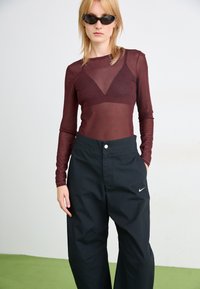 Burgundijas caurspīdīgs ilgs piedurkņu tops ar abstraktiem rakstiem, apvienots ar brīviem melniem biksēm, kuru kreisajā augšstilbā ir balts Nike logo.