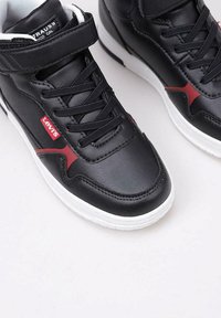 Sneaker alte nere in pelle con suole in gomma bianca. Presentano dettagli rossi e lacci, e una chiusura con strap in Velcro.