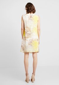 Frau in einem ärmellosen knielangen Kleid mit beige-, gelb- und weißen Palmenblattmuster und beigen Blockabsatzsandalen, mit dem Rücken zur Kamera.