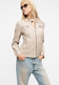 Γυναίκα με μπεζ δερμάτινο μπουφάν με φερμουάρ, ανοιχτό μπλε τζιν και σκουρόχρωμα γυαλιά ηλίου, στέκεται με τα χέρια στις τσέπες μπροστά σε λευκό φόντο.