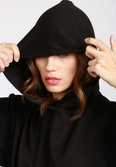 Felpa con cappuccio nera dal design spazioso e cappuccio, caratterizzata da una texture morbida. Il modello si sistema il cappuccio con entrambe le mani, coprendo parzialmente il volto.