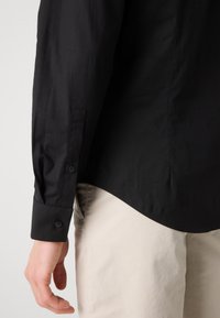 Black long-sleeve shirt with buttoned cuffs, made from smooth fabric. Curved hem and slight sheen. Paired with light beige pants.