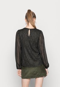 Blusa nera con maniche lunghe e trasparenti, impreziosita da strisce verticali dorate. Presenta un'apertura a chiave sulla schiena e una vestibilità leggermente ampia.