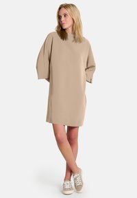 Beige ylipitkä mekko, jossa on korkea kaula-aukko ja lyhyet, leveät hihat. Yhdistetty vaaleisiin tennareihin. Sujuva kangas, polven pituinen siluetti.