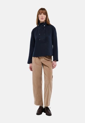 Pull-over bleu marine à col montant, avec deux rangées de boutons et une poche plaquée. Associé à un pantalon beige à jambes larges et des chaussures sombres.