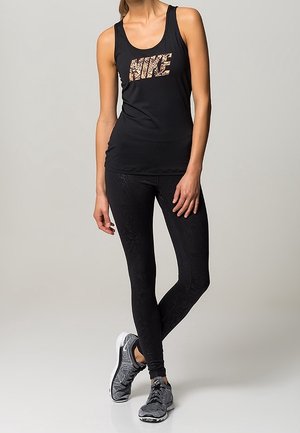 Czarny bezrękawnik z fakturowanym złotym logo Nike, w połączeniu z czarnymi legginsami w wzory i szarymi butami sportowymi.