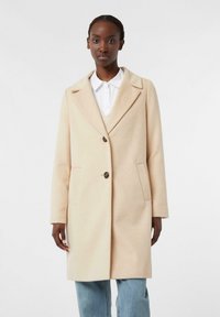 Cappotto in lana beige con rever, due bottoni e tasche laterali. Tessuto liscio e vestibilità su misura, abbinato a una camicia bianca sotto.