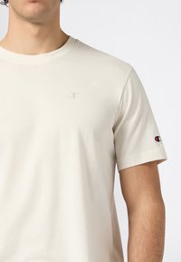 Man in een effen lichtbeige T-shirt met korte mouwen, met kleine Champion-logo's op de borst en de mouw, tegen een witte achtergrond.