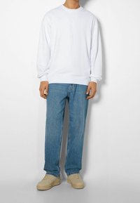 Sudadera blanca de manga larga con puños acanalados, combinada con jeans de corte relajado en azul claro y zapatillas beige. Diseño simple y limpio.