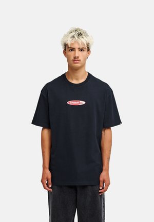 Młody mężczyzna o blond włosach, ubrany w czarną koszulkę Quiksilver i ciemne spodnie, stojący na jednolitym jasnym tle.