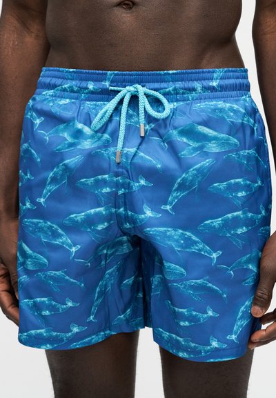 Μπλε μαγιό σορτς με φωτεινά γαλάζια σχέδια φαλαινών και κορδόνι μπροστά, φορεμένα από ένα σκούροχρωμο άτομο από τη μέση μέχρι τη μέση του μηρού.