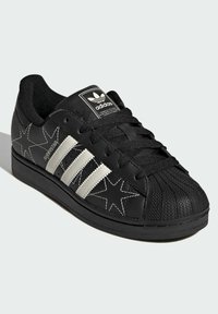 Black Adidas Superstar sneakers with star-shaped stitching, three white stripes on the side, and a textured rubber toe cap.