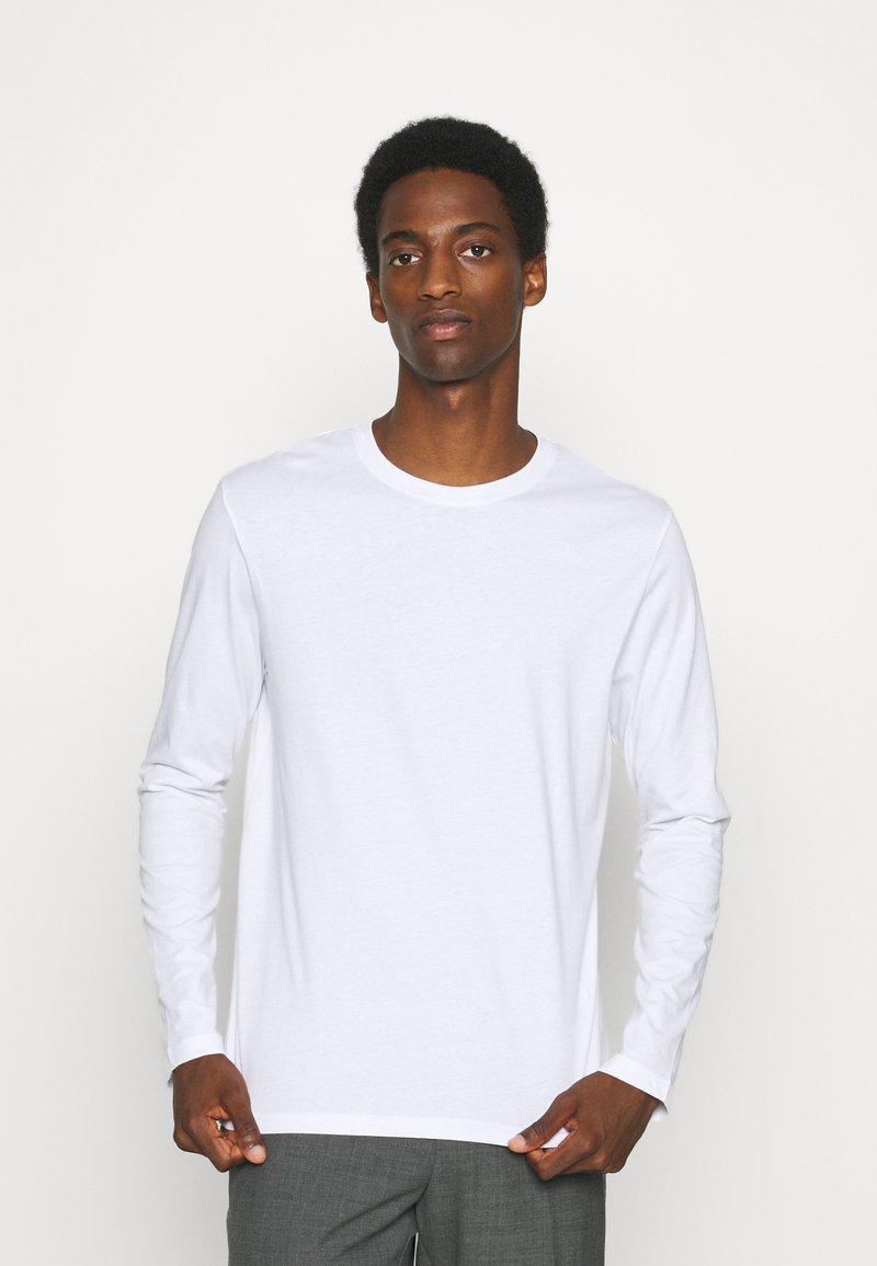 Weißes Langarm-T-Shirt aus Baumwolle, mit Rundhalsausschnitt und schlichter Gestaltung, getragen über grauen Hosen, mit glatter Textur.