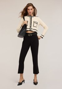 Crème cropped cardigan met zwarte randen en zakken, gecombineerd met zwarte taille broek en puntige pumps, met een zwarte schoudertas.