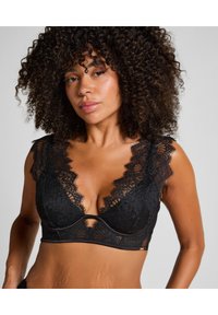 Zwarte kanten bralette met een diepe halslijn en geschulpte afwerking, voorzien van delicate bloemenpatronen en subtiele doorschijnende panelen.