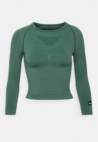 Długi rękaw, zielona bluzka sportowa o prążkowanej teksturze, dopasowany krój, marszczony detal z przodu oraz akcent w postaci czarnej metki na prawym rękawie.
