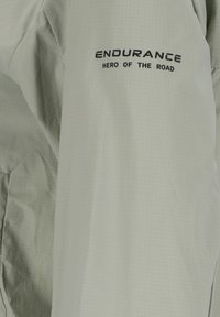 Lys grå cykeljakke med et struktureret gittermønster. Har sort tekst "ENDURANCE HERO OF THE ROAD" på skulderen.