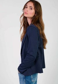 Blazer navy in tessuto morbido con vestibilità slim, chiusura a un bottone e tasche laterali. Indossato sopra una camicia bianca e jeans blu.