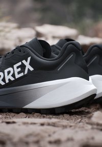 Scarpe sportive bianche e nere con suole spesse su terreno roccioso, che mostrano il lato con un grande testo parziale bianco "ARREX."