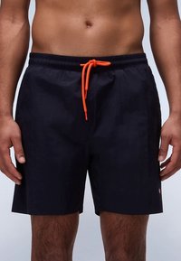 Șort de baie bleumarin cu talie elastică, șnur portocaliu și un detaliu mic de branding pe partea inferioară a piciorului. Material neted și ușor.