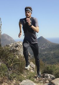 Sportoló futó szürke, rövid ujjú pólóban, fekete, tapadó leggingsben és fekete, világos részletekkel ellátott cipőben, fekete fejpántot és hidratáló hátizsákot visel.