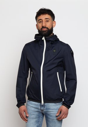 Giacca impermeabile blu navy con zip frontale, due tasche laterali, cappuccio regolabile e dettagli e zip bianchi a contrasto.