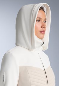 Bijela jakna sa strukturom na kapuljači, koja ima teksturiranu bež ploču. Mekani oblog na vratu i RECCO logo na rukavu.