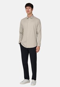 Beige langärmliges Hemd mit Knopfleiste, leichtem Stoff und spitzem Kragen. Kombiniert mit schwarzen Hosen und weißen Turnschuhen.