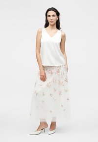 Femme portant un haut blanc sans manches avec une jupe midi blanche transparente brodée de fleurs roses et des chaussures blanches à talons bas.