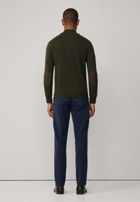 Donkergroene gebreide trui met bruine elleboogpatches, geribde manchetten en kraag. In combinatie met marineblauwe broek en donkerbruine schoenen, gezien van achteren.