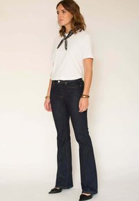 T-shirt blanc à col rond, jean évasé bleu marine avec des coutures jaunes, et ballerines noires pointues, accessoirisées avec une écharpe noire.