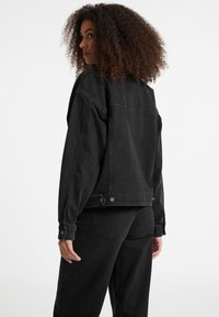 Black denim jacket with a boxy fit, button accents, and visible stitching. Features a classic collar and two back buttons.