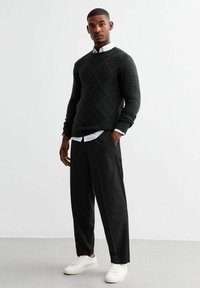 Mörkgrå stickad tröja med diamantmönster, lager-på-lager över en vit krag skjorta, tillsammans med svarta löst sittande byxor och vita sneakers.