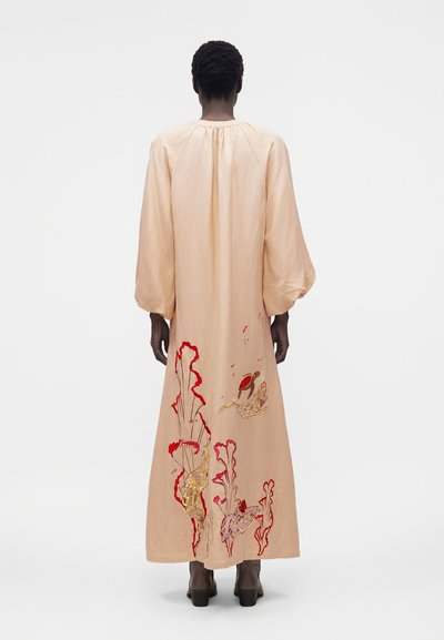 Άτομο που στέκεται με την πλάτη γυρισμένη, φοράει μακρύ μπεζ φόρεμα με κόκκινη, κίτρινη και μοβ κεντητική με λουλούδια και μαύρα μποτάκια μέχρι τον αστράγαλο.