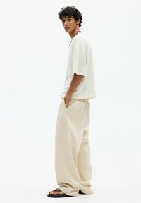 Light beige oversized t-shirt and loose cream trousers with side pockets. Model wears black sandals, showcasing a relaxed casual look.