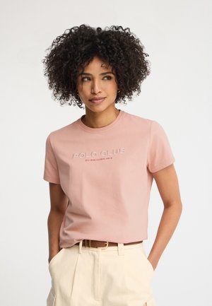 Vrouw met krullend haar die een roze "Polo Club" T-shirt draagt, ingestopt in crème-kleurige broek met hoge taille en een bruine riem, staand met handen in de zakken.