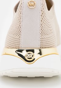 Sneaker in maglia beige con retro strutturato, dotato di una piastra accentuata in oro e dettaglio del logo sul tallone, abbinato a una suola in gomma bianca.