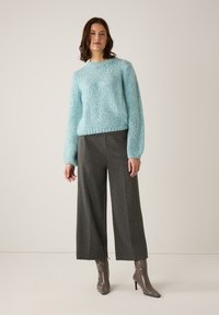 Frau, die einen hellblauen Strickpullover, graue weitgeschnittene, knöchellange Hose und graue, schicke Stiefeletten trägt und vor einem schlichten Hintergrund steht.