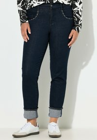 Vaqueros azul oscuro de corte ajustado, con bolsillos decorativos con tachuelas, puños enrollados y una textura de denim suave. Combinados con zapatillas blancas.