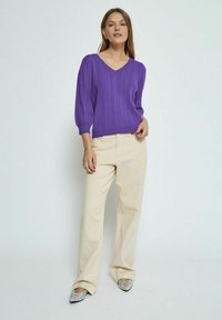 Violet adīta džemperis ar V-veida izgriezumu un teksturētu dizainu, kombinēts ar krēmkrāsas platām biksēm un rotātām smailu degunu kurpēm.