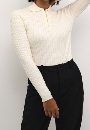 Persoon draagt een crèmekleurige, gebreide trui met lange mouwen, een kraag en een rits, gecombineerd met hoge zwarte broek tegen een effen achtergrond.