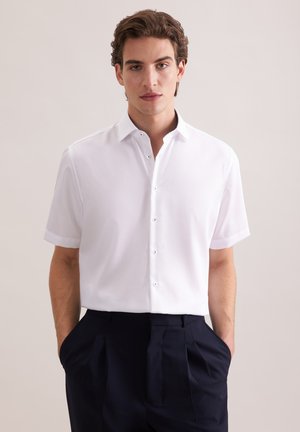 Mężczyzna ubrany w białą koszulę z krótkim rękawem, wpuszczoną w czarne plisowane spodnie, z rękami w kieszeniach, na neutralnym tle.