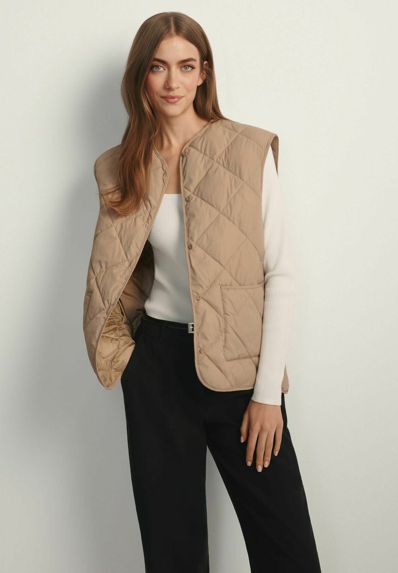 Beżowa pikowana kamizelka z zapięciem na guziki i bocznymi kieszeniami, noszona na białej bluzce z długim rękawem, w zestawieniu z czarnymi spodniami. Gładka faktura materiału.