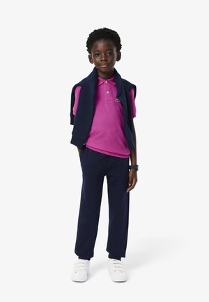 Maglietta polo viola, pantaloni blu navy e sneakers bianche. Maglione blu navy appoggiato sulle spalle. Bambino in piedi con sicurezza su uno sfondo neutro.
