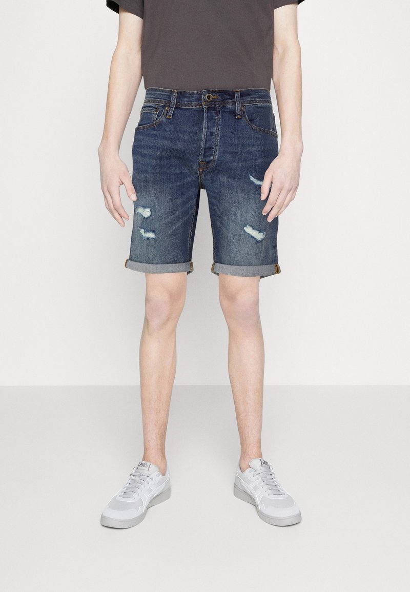 Jeansshorts i en mörkblå tvätt, med slitna detaljer, uppvikta benslut och femficksdesign. Kombinerade med grå sneakers.