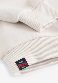 Světle béžová mikina s žebrovanými manžetami. Má jemnou texturu a na manžetě je látková visačka se známkou "SUPERDRY" v červené a modré barvě.
