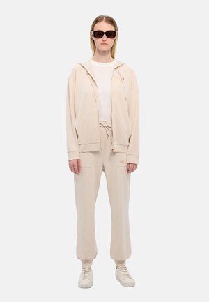 Felpa beige con zip e cordino, abbinata a pantaloni jogger coordinati. Sotto indossa una maglietta bianca semplice, con scarpe da ginnastica bianche.