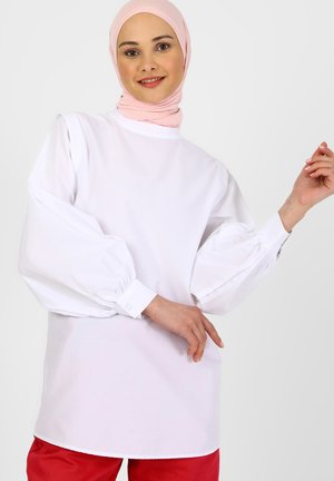 White long-sleeve top with puffed sleeves and buttoned cuffs, paired with red pants. Smooth texture, high neckline, and a relaxed fit.